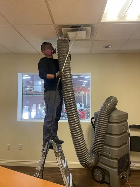 Mighty Ducts technician cleaning a ceiling air duct with a commercial duct-cleaning vacuum hose in an office